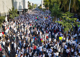 Una gran multitud de personas se reunió en una calle, muchas de ellas con camisetas blancas, con árboles y edificios de fondo. Algunos asistentes portaban carteles de colores.