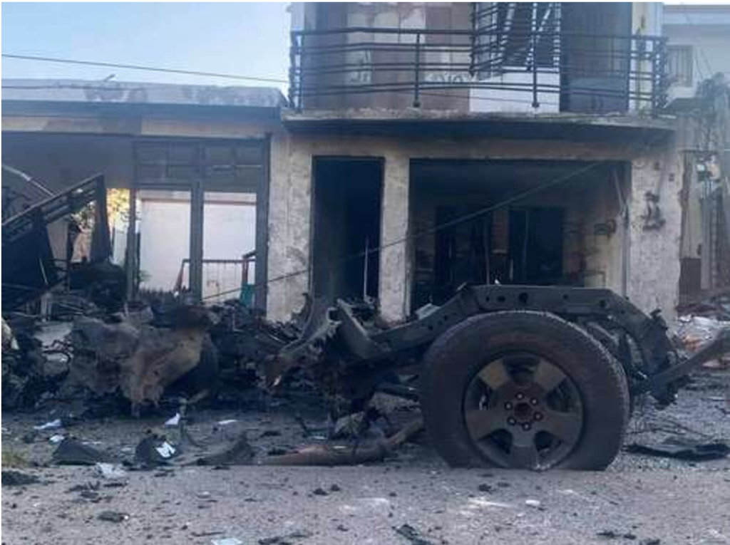 Restos carbonizados de un vehÃ­culo frente a un edificio daÃ±ado, con escombros esparcidos en el suelo.