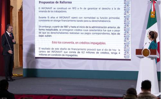 Dos personas están de pie en un escenario presentando propuestas de reforma proyectadas, con texto en español en una pantalla grande. Se ve parcialmente una bandera mexicana en el fondo.