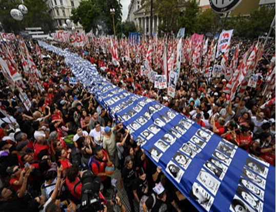 Una gran multitud marcha con pancartas, sosteniendo una larga tela azul que muestra fotografías en blanco y negro de personas, rodeadas de numerosas banderas rojas y blancas.