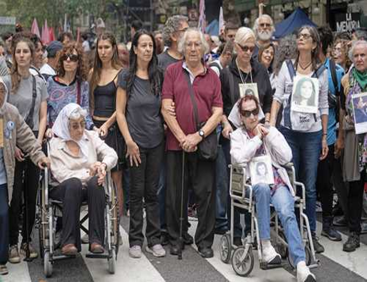 Un grupo diverso de personas, incluyendo personas en silla de ruedas, participa en una reunión al aire libre, algunas con retratos. El fondo muestra una concurrida calle de la ciudad.