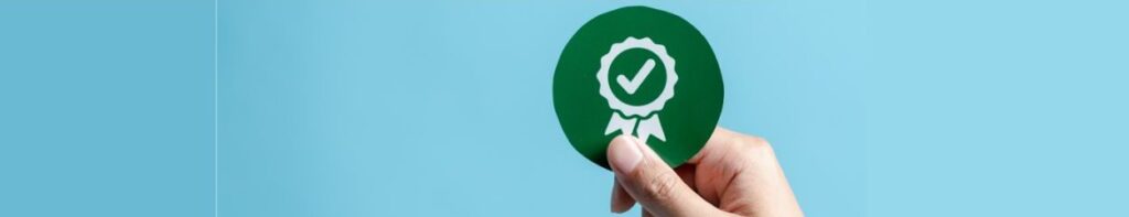 Una mano sostiene una insignia verde con un sÃ­mbolo de marca de verificaciÃ³n sobre un fondo azul claro, que significa Cumplimiento Aduanero.