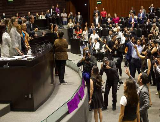 Multitud de personas dentro de una gran sala, algunos tomando fotos, otros gesticulando, cerca de una plataforma elevada con individuos sentados.