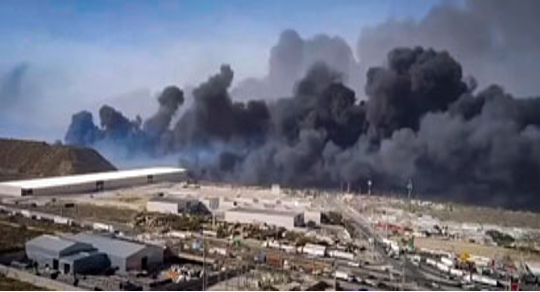 Una gran zona industrial está rodeada de un denso humo negro que se eleva hacia el cielo, lo que indica un incendio importante.