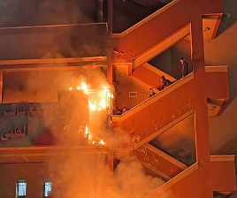 Incendio en la escalera exterior de un edificio con llamas visibles. Varias personas permanecen en la escalera, con aire cauteloso.