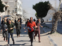 Un grupo de personas corre por una calle llena de edificios dañados. Un hombre con camisa roja carga a un niño. Se ven polvo y escombros al fondo.