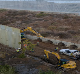 Los trabajadores de la construcción y la maquinaria excavan cerca de una barrera y de tuberías de drenaje. Un vehículo blanco está estacionado cerca.