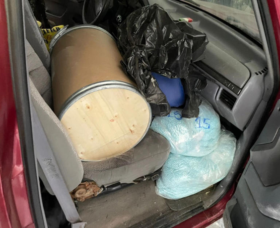 Interior del automÃ³vil con barril de madera en el asiento trasero, bolsas de plÃ¡stico negras y bolsas azules grandes etiquetadas con texto blanco.