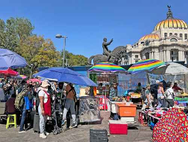 La gente pasea por un mercado con sombrillas de colores cerca de un edificio abovedado y una estatua ecuestre bajo un cielo azul claro.