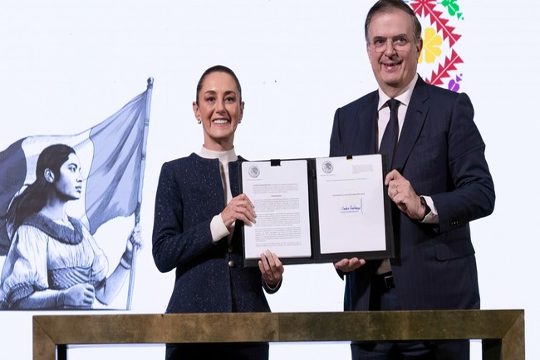 Dos personas sostienen un documento firmado. Al fondo se ve un mural de una mujer con una bandera.