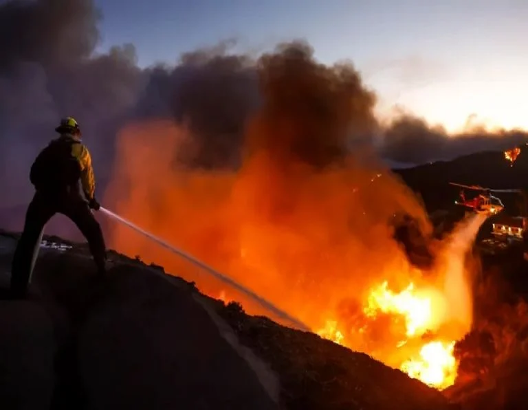 Un bombero rocía agua sobre un gran incendio forestal bajo un cielo oscuro y lleno de humo, con un helicóptero visible cerca.