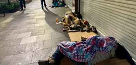 Una persona cubierta con una manta colorida yace en una acera cerca de una tienda, con otra figura y pertenencias en el fondo.
