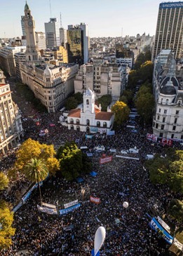 Vista aÃ©rea de una gran multitud reunida alrededor de una pequeÃ±a iglesia blanca en una plaza urbana, con edificios circundantes y pancartas visibles.