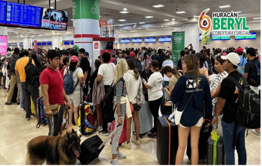 Una terminal de aeropuerto abarrotada, con numerosos pasajeros haciendo cola con equipaje. En lo alto se ven pantallas que muestran diversa información de vuelos. La zona está muy concurrida y también hay un perro entre los viajeros.