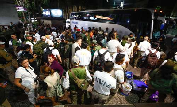 Un gran grupo de personas, entre ellos uniformados y civiles, se reÃºnen por la noche cerca de dos autobuses blancos. Algunas personas llevan mochilas y equipo, mientras que otras conversan.