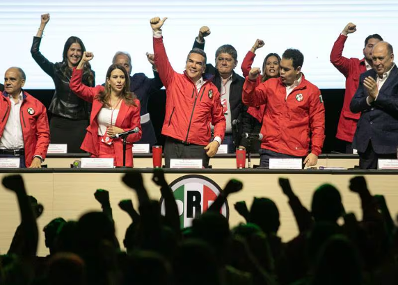 Un grupo de personas, en su mayorÃ­a vestidas con chaquetas rojas, estÃ¡n de pie en un escenario, algunas levantando los puÃ±os. En primer plano hay una multitud vitoreando con los puÃ±os en alto. En la mesa frente a ellos se ve el logotipo del "PRI".