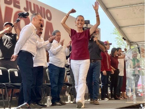 Una mujer con un top rojo y jeans levanta los brazos triunfalmente en el escenario de un evento público, rodeada de hombres que aplauden.
