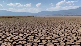 Un primer plano de tierra seca y agrietada con un lago y montañas a lo lejos bajo un cielo despejado.