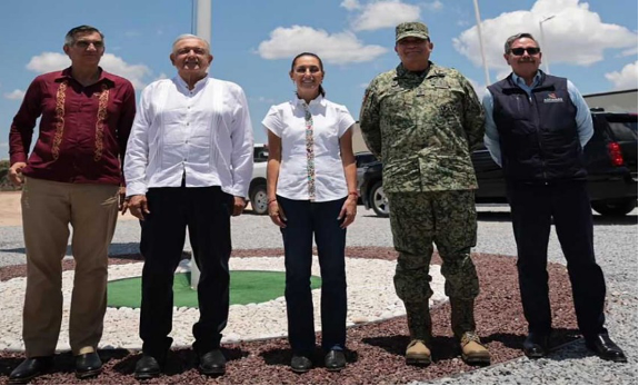 Cinco personas se encuentran al aire libre sobre una superficie de grava, incluidos dos hombres con camisas tradicionales, una mujer con una blusa blanca, un hombre con uniforme militar y otro con chaqueta y gafas de sol.