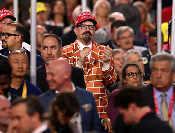 Un hombre vestido con un traje diseÃ±ado para parecerse a una pared de ladrillos y un sombrero rojo que dice "Make America Great Again" aplaude entre la multitud en un evento.