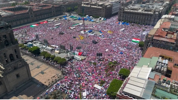 Vista aérea de una gran multitud que llena una plaza de la ciudad, con gente vestida de diferentes colores y algunas banderas visibles. Los edificios bordean la plaza y se instalan varias tiendas de campaña y escenarios en medio de la multitud.