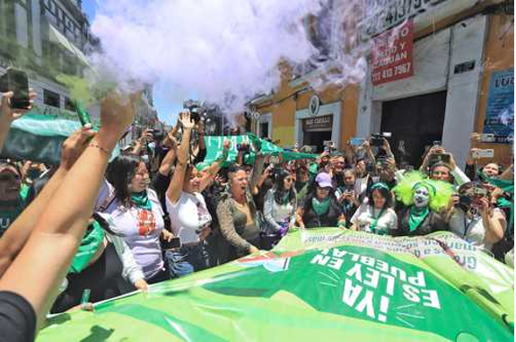 Un grupo de personas con pancartas verdes y pañuelos se reúne en una calle, algunos levantan sus teléfonos para tomar fotos o vídeos mientras el humo se eleva en el aire.