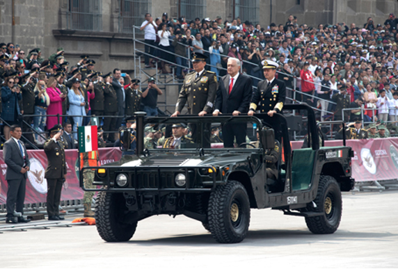 Tres hombres vestidos con uniforme militar de gala se encuentran de pie en un vehículo verde durante un desfile, con una gran multitud al fondo. Una bandera mexicana está colocada en la parte delantera del vehículo.