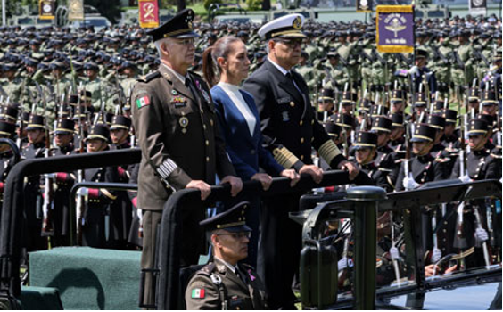 Oficiales militares y una mujer se encuentran en un vehículo abierto durante un gran desfile militar, con filas de soldados uniformados alineados detrás de ellos.
