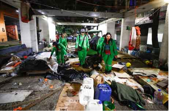 Un grupo de personas con uniformes verdes inspeccionan un refugio improvisado y abarrotado de objetos desechados y escombros debajo de una estructura de concreto.