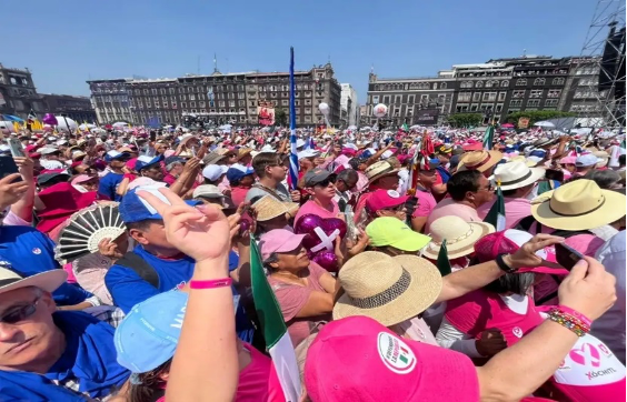 Una gran reunión de personas vestidas de colores, incluidos rosa y azul, sosteniendo carteles y banderas en una plaza pública con edificios antiguos al fondo.