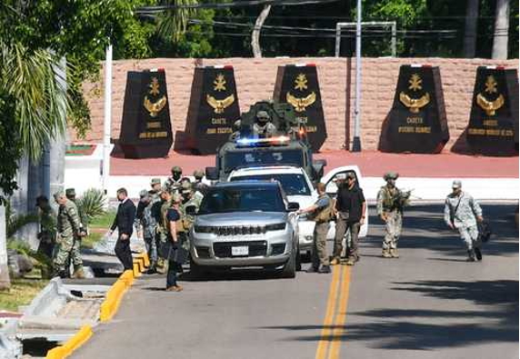 Personal militar y vehÃculos se encuentran estacionados en una carretera cerca de un monumento con placas y emblemas. Al fondo se ven árboles y un muro de ladrillos.