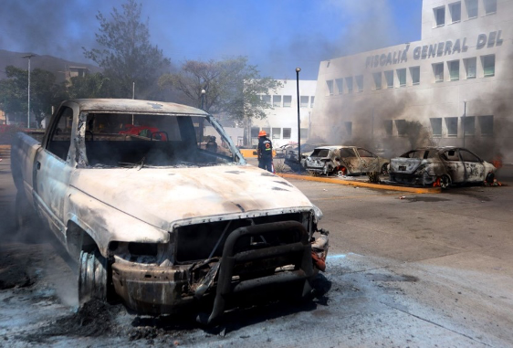 Bombero caminando cerca de vehÃculos quemados con humo de fondo.