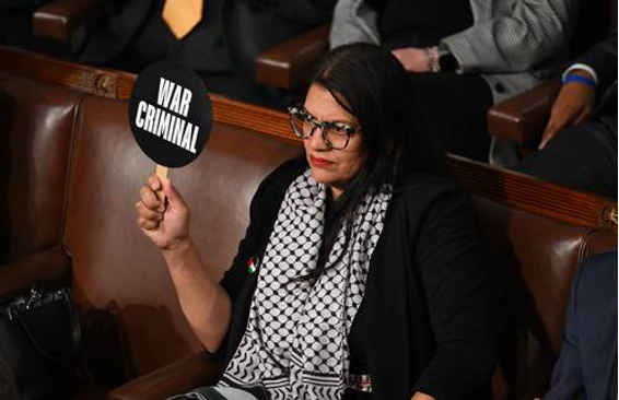 Persona sentada, sosteniendo un cartel que dice "CRIMINAL DE GUERRA", con gafas y un pañuelo estampado alrededor del cuello.