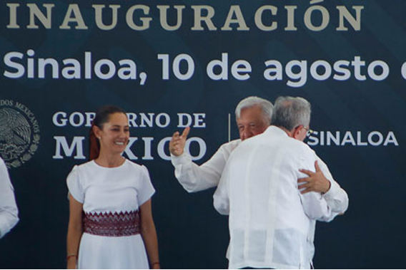 Varias personas se encuentran en el escenario durante un evento en Sinaloa, MÃ©xico. Un hombre en el fondo hace un gesto con el pulgar hacia arriba mientras otro hombre en primer plano recibe un abrazo. Una mujer estÃ¡ de pie a la izquierda, sonriendo.
