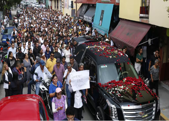 Una gran multitud sigue a un vehículo negro cubierto de pétalos de flores rojas y blancas. Una persona al frente sostiene un cartel que dice "Queremos paz en Chiapas".
