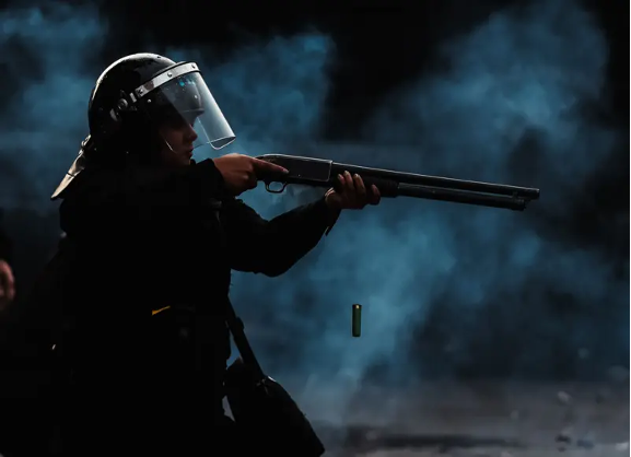Una persona que lleva un casco con visera apunta con una escopeta en un ambiente oscuro y lleno de humo, con un casquillo de escopeta visiblemente expulsado en el aire.