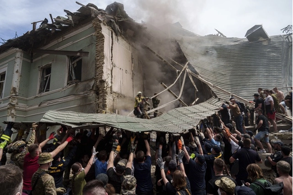 Un grupo de personas, incluidos rescatistas y civiles, trabajan juntos para levantar los escombros de un edificio parcialmente derrumbado del que se eleva humo, probablemente durante una operación de rescate después de un desastre.