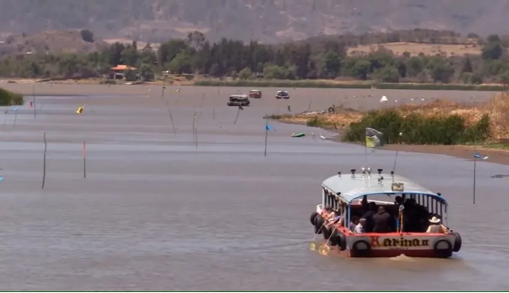Un barco con pasajeros navega por un lago tranquilo con colinas distantes y escasa vegetaciÃ³n visible al fondo.
