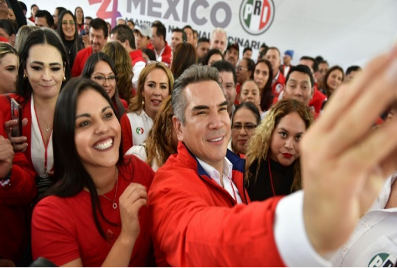 Un grupo de personas, en su mayoría vestidas de rojo, se reúnen y sonríen para tomarse una selfie. Un hombre en el centro extiende el brazo sosteniendo el teléfono para tomar la foto. Una pancarta al fondo dice "México PRI".
