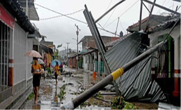 Varias personas caminan por una calle inundada con escombros y estructuras derrumbadas después de una tormenta. Una persona sostiene un paraguas mientras otra lleva una bolsa.