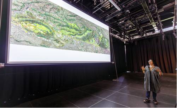 Una persona se encuentra frente a una gran pantalla de proyecciÃ³n que muestra un mapa topogrÃ¡fico detallado en una habitaciÃ³n oscura con cortinas negras.