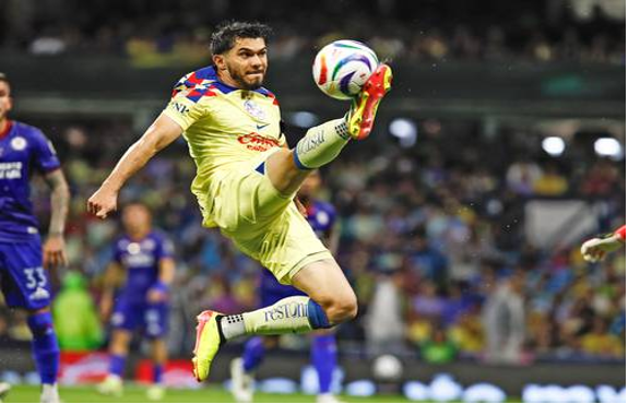 Un jugador de fÃºtbol con uniforme amarillo salta para patear el balÃ³n en el aire durante un partido, con los espectadores y otro jugador de azul al fondo.