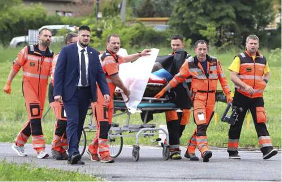 Los socorristas vestidos de naranja llevan a una persona en una camilla. Un hombre con traje azul camina junto a ellos. La escena se desarrolla al aire libre, con hierba y árboles visibles al fondo.