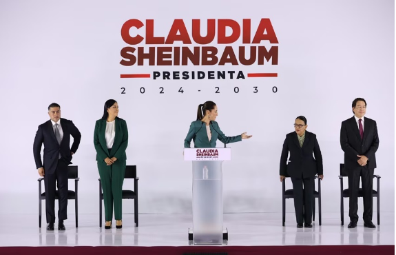 Cinco personas estÃ¡n en el escenario, con una mujer hablando en un podio con la leyenda "Claudia Sheinbaum, Presidenta 2024-2030". Un gran telÃ³n de fondo muestra su nombre y tÃ­tulo.