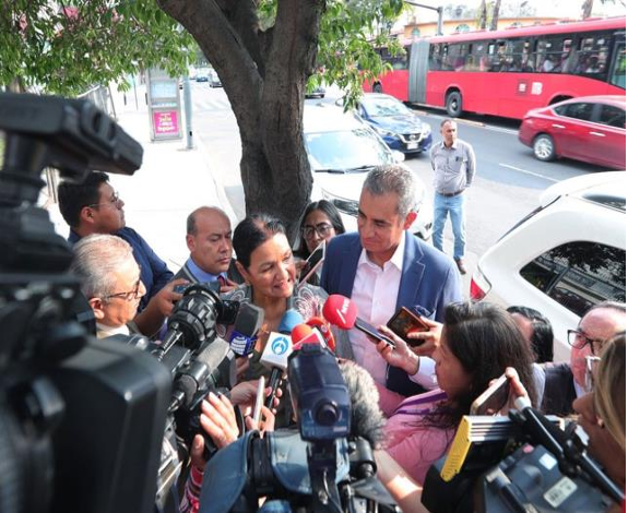 Un grupo de periodistas con micrófonos y cámaras rodea a dos personas sonrientes cerca de un árbol en una concurrida calle de la ciudad con un autobús rojo al fondo.