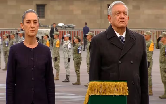 Dos personas se encuentran de pie solemnemente en un evento al aire libre, con soldados uniformados en formación detrás de ellas. En primer plano se ve parcialmente un podio cubierto con flecos.