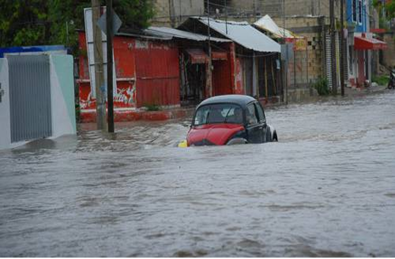Un coche rojo y negro está parcialmente sumergido en una calle inundada llena de edificios, tras una fuerte lluvia.