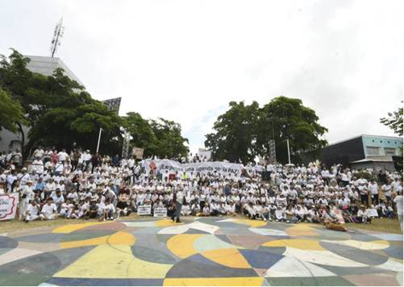 Un gran grupo de personas, en su mayoría vestidas de blanco, se reúne al aire libre frente a árboles y edificios, sosteniendo una pancarta que aboga por la paz. El suelo muestra un patrón abstracto y colorido.