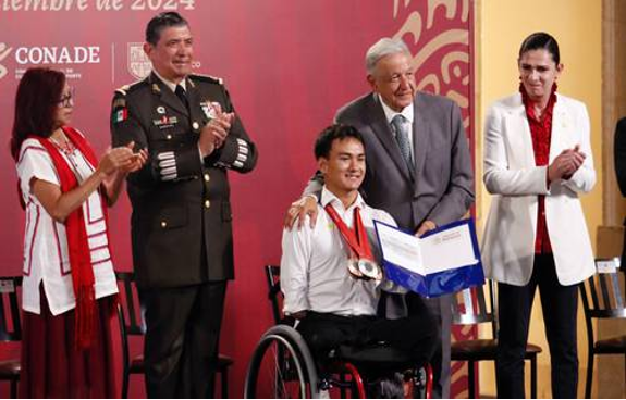 Un hombre en silla de ruedas recibe un premio de manos de un hombre mayor vestido de traje. Otras tres personas, entre ellas un oficial militar, están de pie junto a ellos aplaudiendo.