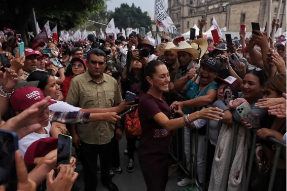 Una mujer vestida de granate saluda a una multitud; algunos sostienen teléfonos inteligentes y usan sombreros, incluidos sombreros de vaquero, mientras que otros toman fotografÃas. Al fondo se ven un edificio y árboles.